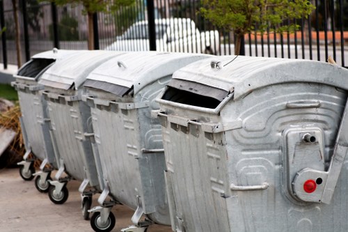 A row of four large metal wheelie bins lined up on a paved surface, with the front of the bins facing the camera. The bins are made of galvanized steel with a reflective, slightly textured finish and feature black plastic lids that are partially open. Each bin has four black wheels at the base for mobility, positioned on a concrete or tarmac area likely outdoors. Behind the bins, a black metal fence and some leafy green trees are visible, suggesting a residential or commercial area in Penge. The scene appears to be captured in daylight, with soft natural lighting. The image reflects the typical outdoor rubbish and waste management setup that a professional rubbish removal service like Garden Clearance Penge might handle, focusing on waste receptacles used for general waste or garden clearance rubbish.