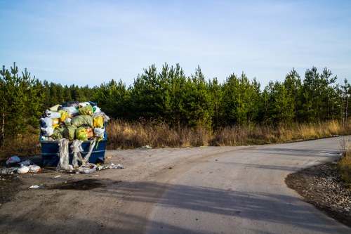 The image shows a large blue skip filled with various types of rubbish, including plastic bags, cardboard boxes, and other waste materials, situated on the side of a rural road. The skip is overflowing with dumped waste, some of which is spilling onto the gravel surface beneath it, with additional rubbish scattered around its base. In the background, there is a line of dense green trees under a clear blue sky, indicating a natural environment surrounding the roadside. The scene suggests an area where waste has been disposed of improperly, highlighting the importance of proper rubbish removal services. The setting appears quiet, with no visible people or vehicles, and the road curves to the right, leading away from the scene. This visual context aligns with the type of clutter that Garden Clearance Penge might help remove from gardens, driveways, or residential areas, emphasizing the need for professional outdoor waste removal in the local area near Penge and the surrounding South London region.